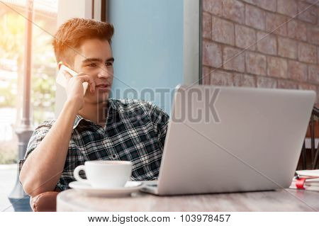 Businessman Using Mobile Phone While Lookingat Laptop On Wooden Table In Coffee Shop