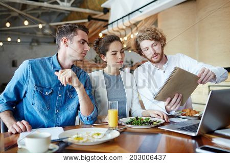 Clever student showing his sketch for coursework in notepad to groupmates during lunch break in cafe