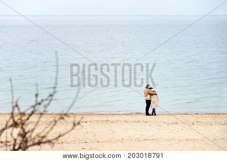 Happy loving couple embracing wearing warm clothes standing on ocean coast. Cold season, sea shore, empty beach in autumn or winter