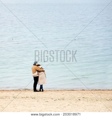 Happy loving couple embracing wearing warm clothes standing on ocean coast. Cold season, sea shore, empty beach in autumn or winter