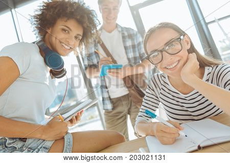 Happy team. Close up of positive smiling young international students sitting at the table and making notes while enjoying studying together