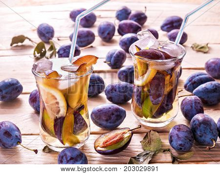 Refreshing drink from fresh juicy sweet plums with lemon in glass mugs with ice cubes on a wooden background with plums and green leaves