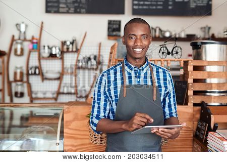 Portrait of a smiling young African entrepreneur in an apron leaning against the counter of his trendy cafe using a digital tablet