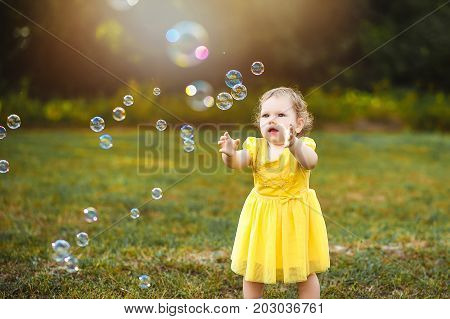A little girl in a yellow dress in a green glade is catching soap bubbles.