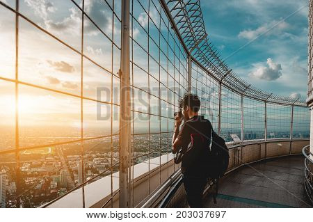 Man photographer in black dress taking pictures of buildings on the top floor of the building Baiyoke Tower Bangkok Thailand during the twilight sun. Light is very beautiful