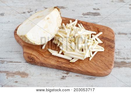 Root celery cut into small pieces with the half of the whole celery root on a olive wood cutting board on a light wooden background. Step by step cooking.