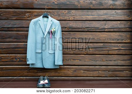 Stylish elegant wedding groom suit with buttonhole hanging on wooden background with copy space. Gray suit hangs above leather groom shoes and pink bowtie. Groom wedding accessories free space