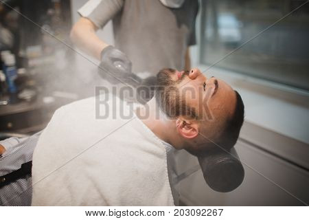 A man who sitting in a barber chair getting beard haircut, a professional barber using a talcum powder on a light blurred background. Hairstyle, haircut, barbershop concept.