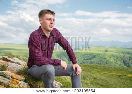 A young guy clasically dressed sits on a rock in nature enjoying a view of the background of Caucasian beauties
