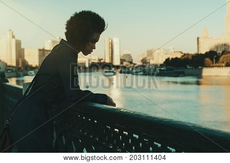Silhouette of young curly black girl leaning on metal fencing of embankment and looking down on the river water while having rest during walking in Moscow; with copy space area for logo or your text
