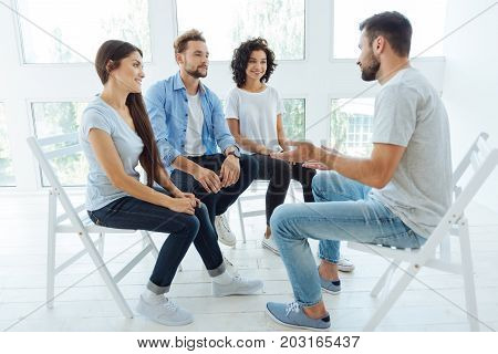 Teambuilding coach. Handsome positive nice man sitting in front of his group and talking to them while working as a teambuilding coach