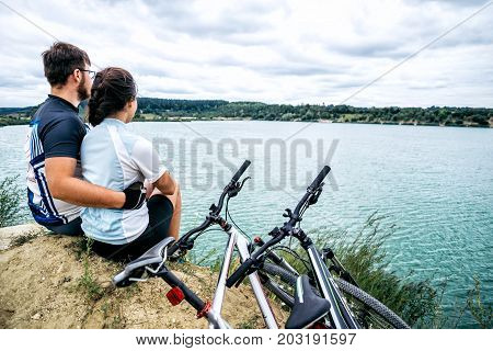 couple sits and looking at the lake two bicycles near them zure water on background