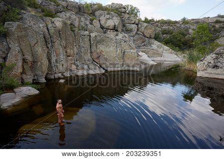A full-length portrait of a muscular swimmer standing in increadibly beautiful pond in abandoned granite quarry. A man on a picturesque canyon background. Nature, summer, travel concept. Copy space.