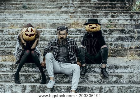 Halloween people or friends on stony stairs. Women with pumpkin heads. Bearded man sitting with girls. Halloween jack o lantern symbols. Holiday celebration concept.
