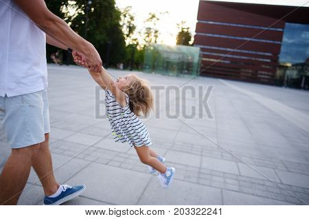Young dad is playing with a charming little daughter. The man holds the baby by hands and is turned together with her. The child rejoices.