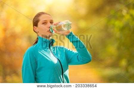 Young woman drinking fresh water from bottle during her fitness exercise in autumn. Lifestyle and sport photo of healthy style. Outdoor and nature fitness.