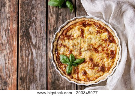 Baked homemade quiche pie in white ceramic form served with fresh greens and textile towel on old wooden plank background. Flat lay with copy space