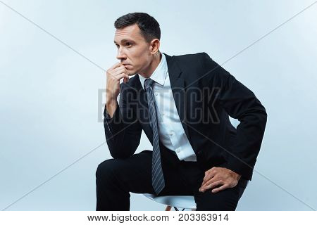 Thoughtful pose. Nice thoughtful smart man sitting on the chair and holding his chin while thinking about his work