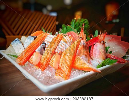 Close-up of mixed sashimi set (Japanese food) on white plate; raw fishes, shrimp, surimi (imitation crab meat) on ice to keep fresh, decorate with green vegetables - Selective focus.