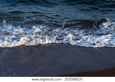 Clear sea water tide on dark volcanic beach. Relaxing sea wave surf over seashore. Coastal seaview. Seascape for summer vacation. Exotic island seaside banner. Sea close-up. Seawater splash on beach