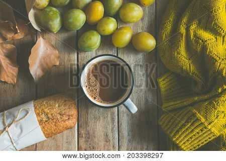 Cup of hot chocolate or cocoa whole grain rye bun scattered yellow and green plums in craft paper bag. Dry leaves knitted woolen sweater on plank wood background. Autumn fall top view cozy.