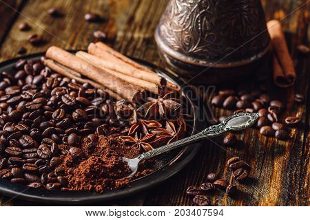 Coffee Beans with Spooonful of Ground Coffee Cinnamon Sticks and Chinese Star Anise on Metal Plate. Beans Scattered on Wooden Table and Cezve on Backdrop.