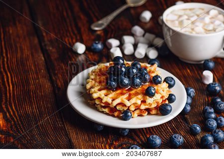 Belgian Waffles on Plate with Fresh Blueberry and Cup of Hot Cocoa with Marshmallow. Spoon and Some Berries and Marshmallows Scattered on Wooden Surface.