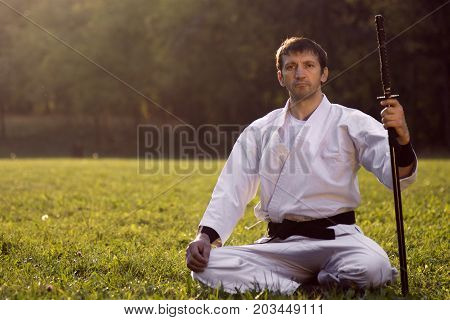 The expert of martial arts is sitting in white kimono with sword on grass outdoor.