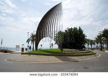 CAMBRILS, SPAIN - AUG 27th, 2017: Modern sculpture A tota vela, a round about and the main street promenade in the port area with Cambrils sign in the background.