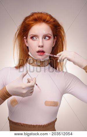 Beautiful Woman before Plastic Surgery Operation Cosmetology. Beauty Portrait Fashion Concept. Treatment with botox or hyaluronic collagen injection. Young red hair girl holding syringes in hands pointing on her lips and breast. Scared female face with co