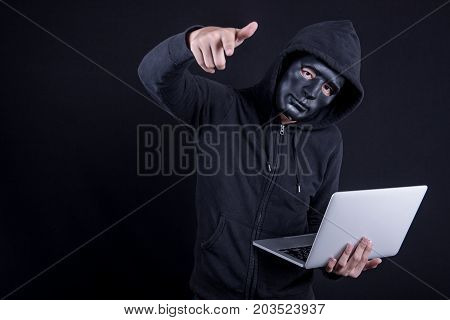 Anonymous male hacker using black mask to cover his face carrying laptop computer and pointing his hand. Internet security and cyber attack concepts.