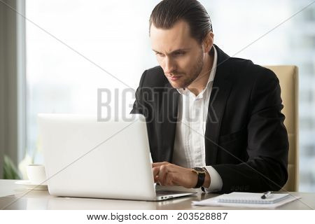 Serious young handsome busy businessman working on laptop at desk at workplace in office. Researching stock investment market, communicating with business partners, taking care of financial inquiries.