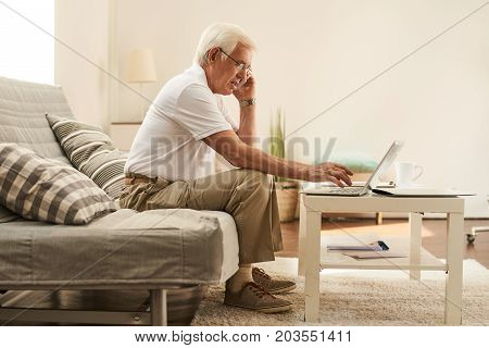 Side view portrait of modern senior man using laptop at home and speaking by phone