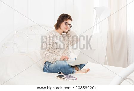 Young woman freelancer works at home in bed with laptop. Engrossed Girl in eyeglasses. Freelance.