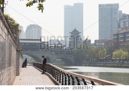 Chengdu, Sichuan Province, China - March 27, 2017: Man watching his smartphone by the jinJinag river with Anshun bridge in the background.