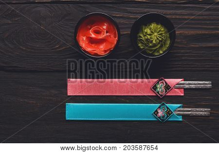 Pieces of traditional japanese tablewear. Chopsticks in blue and pink cases, small black bowls with ginger and wasabi on dark wooden table. Top view, flat lay, copy space, background