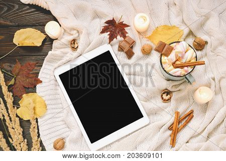 Autumn frame made of dried fall leaves, mug of cocoa with marshmellows, nuts, cinnamon, plaid, apples with tablet mock up. Top view on brown wood background. flat lay, copy space