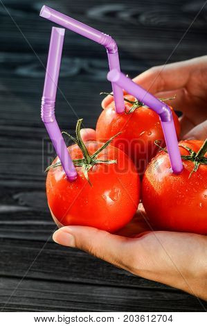 Three Ripe Red Tomatoes with Straws in Hands on Black Wooden Background. Fresh Juice Healthy Food Concept. Tomato as a Vessel with Fresh Juice