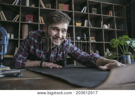 Favorite work in workshop. Young leather maker start work with smile on face and keep leather on wooden table. Exclusive leather product with handmade studio