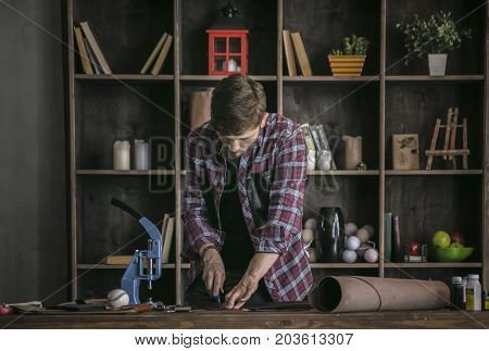 Hard work. Young man leather manufacturer working standing at wooden table and cutting piece of leather for sewing new product. Handmade small business at home