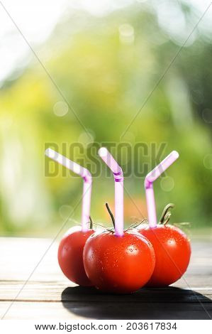Three ripe red tomatoes with straws on black wood table on blurry green background. Fresh juice healthy food concept. Tomato as a vessel with fresh juice