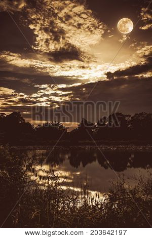 Sky With Dark Cloudy And Full Moon Above Silhouettes Of Trees And Tranquil Lake.