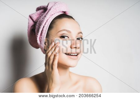 A woman, after a shower with a towel on her head, takes pleasure in caring for the skin, massaging her cheeks.