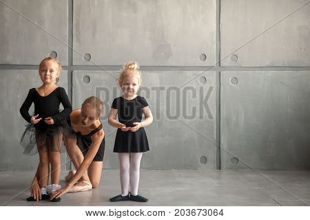 Young beautiful blonde woman ballet dancer in black dress bulging pantyhose and pointe shoes tying pointes to small fair-haired girls ballerines in black dresses in a dark dance studio