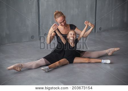 A beautiful adult blonde Russian ballerina in a black tutu and white pantyhose holds her hands up sits on a string and teaches a charming little blonde how to properly dance a Russian ballet