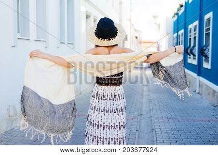 A young woman is walking along a narrow street with a hat. Back view