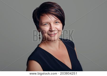 Closeup portrait of beautiful smiling middle aged white caucasian brunette woman with blue eyes in black dress looking in camera on light background in studio