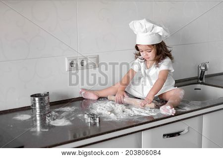 Little smiling baby girl baker in white cook hat and apron kneads a dough on tle kitchen.