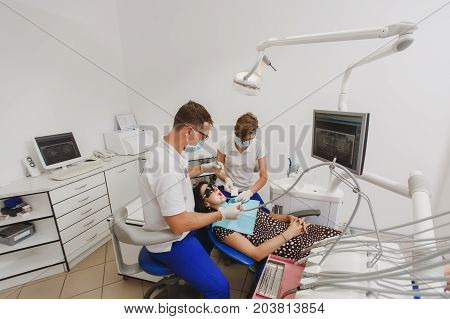 A Male Dentist With A Female Assistant Helps To Treat The Teeth Of A Woman Patient In A Clinic In Th