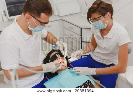 A Male Dentist With A Female Assistant Helps To Treat The Teeth Of A Woman Patient In A Clinic In Th